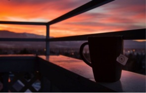 Photo shows a darkly shadowed mug of tea sitting on a railing with a bright orange sunset in the distance